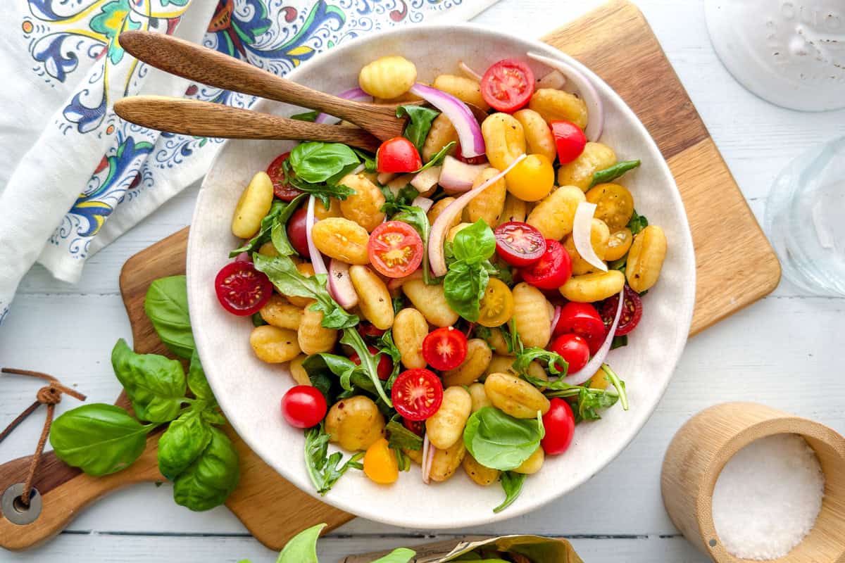 A bowl of gnocchi salad with cherry tomatoes, red onion, fresh spinach, and basil, served on a wooden board with a fork and spoon; colorful napkin and salt container nearby.