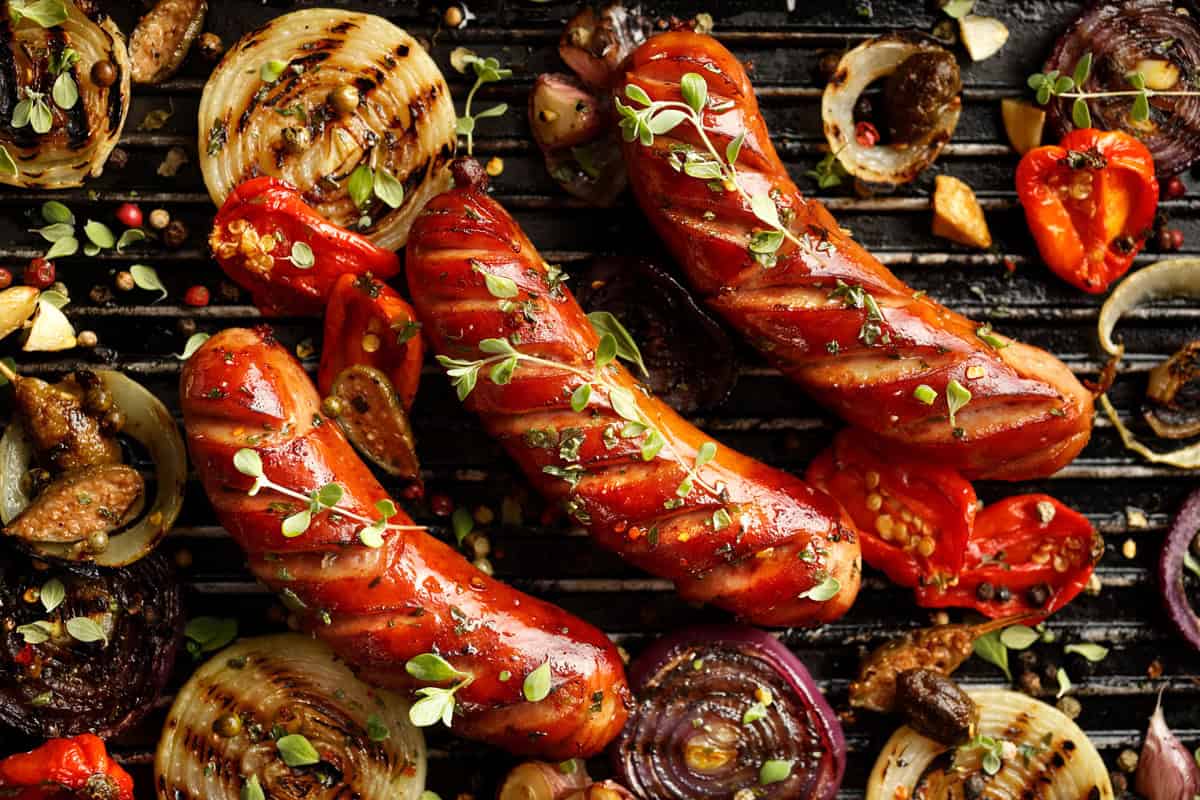 Three grilled sausages with grill marks are topped with herbs and surrounded by grilled onions, tomatoes, garlic, and spices on a black grill surface.