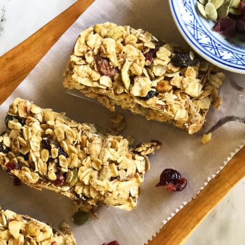 Three homemade granola bars with oats, nuts, and dried fruit rest on parchment paper atop a wooden tray. A blue and white bowl with seeds and cranberries sits in the corner.