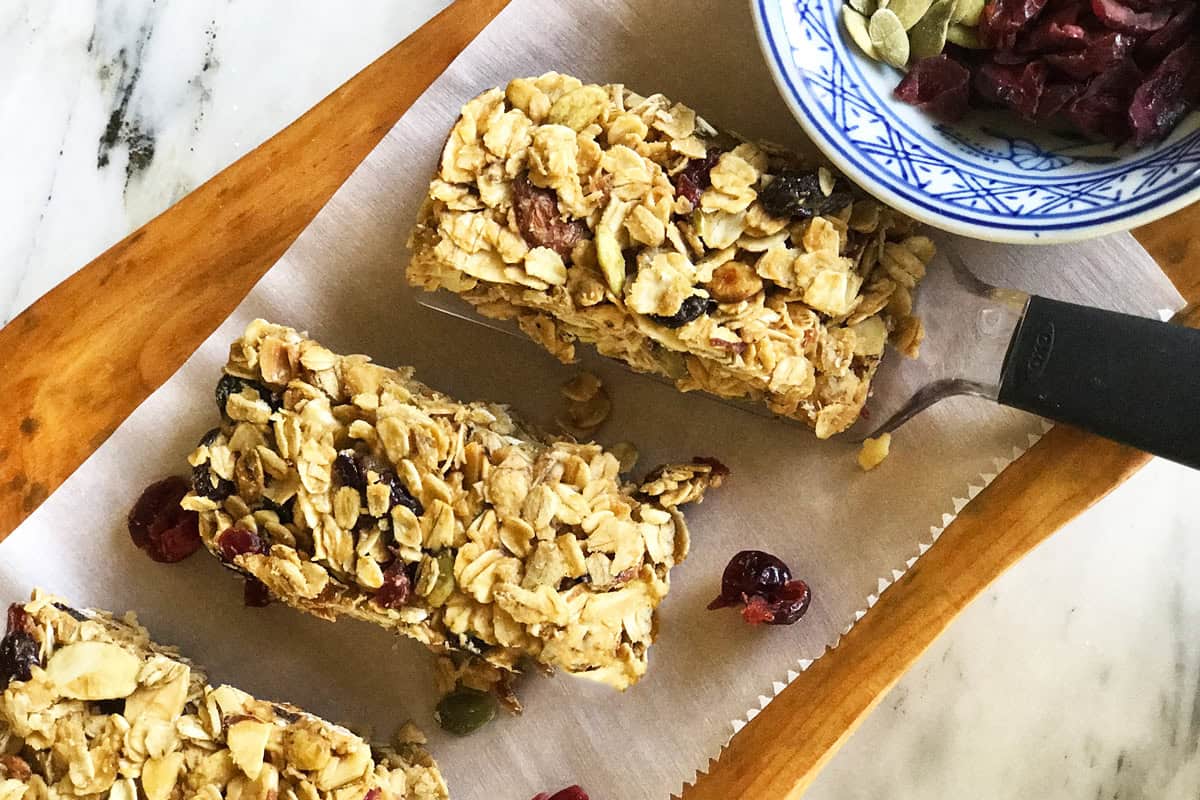 Three homemade granola bars with oats, nuts, and dried fruit rest on parchment paper atop a wooden tray. A blue and white bowl with seeds and cranberries sits in the corner.