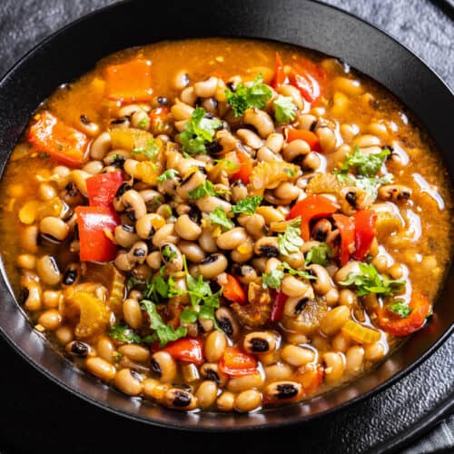 A black bowl filled with black-eyed pea stew featuring chopped tomatoes, bell peppers, celery, herbs, and a savory broth, with a silver spoon beside it on a dark plate.