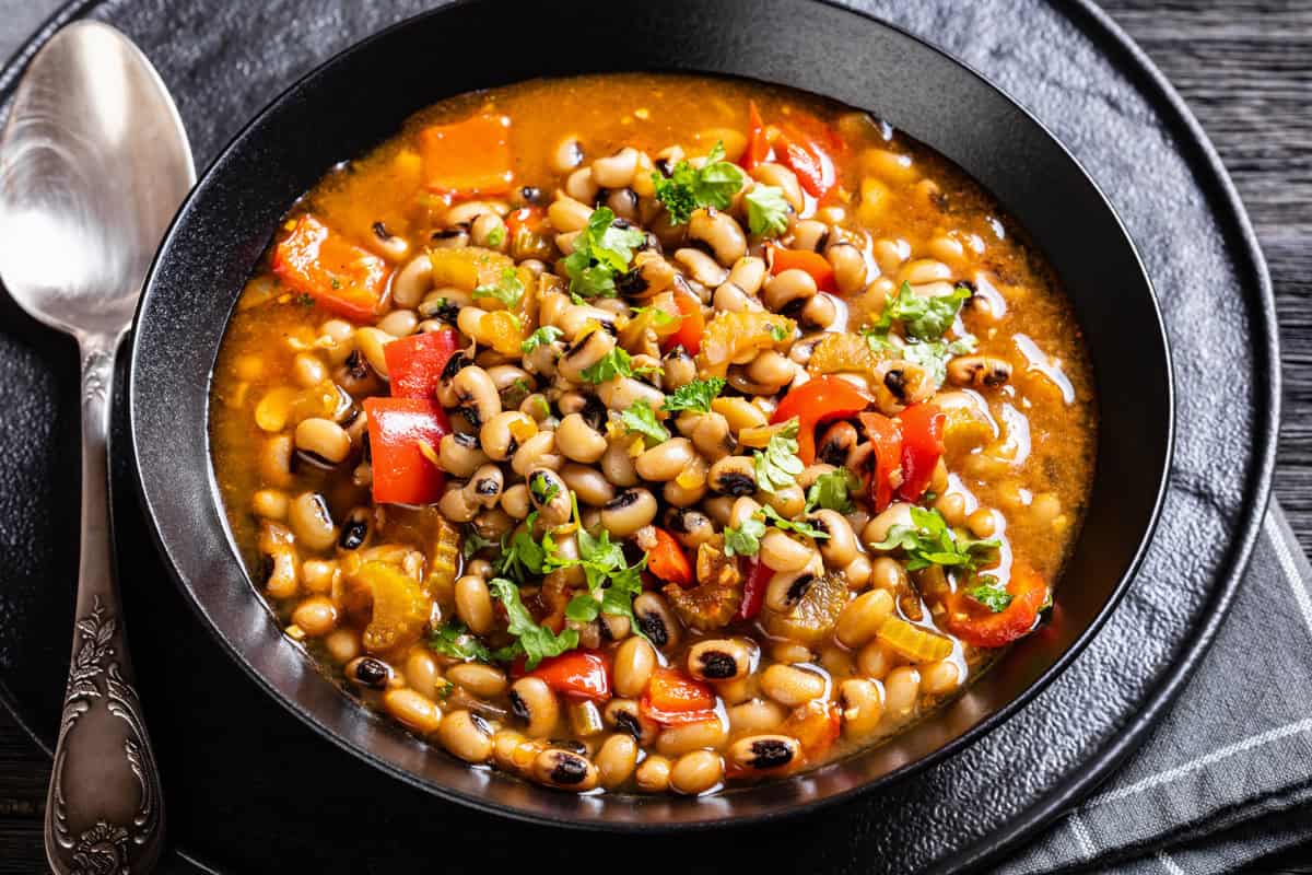 A black bowl filled with black-eyed pea stew featuring chopped tomatoes, bell peppers, celery, herbs, and a savory broth, with a silver spoon beside it on a dark plate.