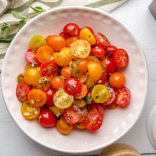 A white bowl filled with sliced red, yellow, and orange cherry tomatoes, garnished with chopped herbs, sits on a light wooden table next to a garlic bulb and a striped cloth.