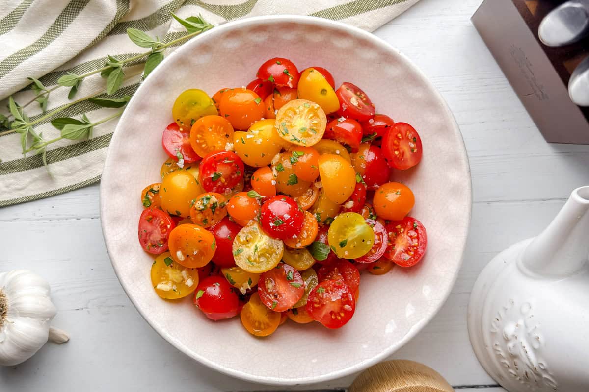 A white bowl filled with sliced red, yellow, and orange cherry tomatoes, garnished with chopped herbs, sits on a light wooden table next to a garlic bulb and a striped cloth.