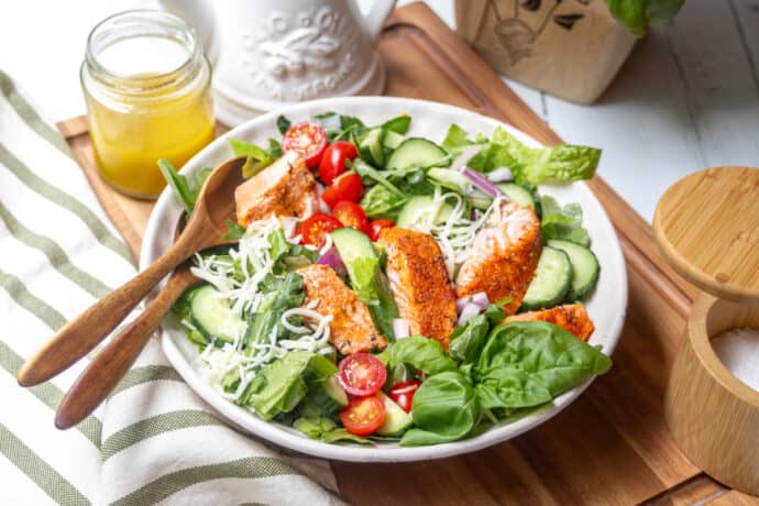 A fresh salad with grilled salmon strips, cherry tomatoes, cucumbers, spinach, shredded cheese, and red onions is served in a white bowl with wooden utensils. A jar of dressing and a salt container are nearby on a wooden board.