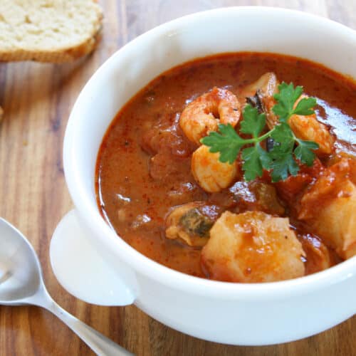 A white bowl of seafood stew, garnished with fresh parsley, sits on a wooden table. A silver spoon and two slices of bread are placed beside the bowl.