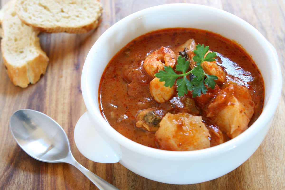 A white bowl of seafood stew, garnished with fresh parsley, sits on a wooden table. A silver spoon and two slices of bread are placed beside the bowl.