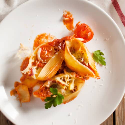 A white plate with stuffed pasta shells in tomato sauce, topped with grated cheese and garnished with parsley, sits on a wooden table next to a white napkin with a red stripe.
