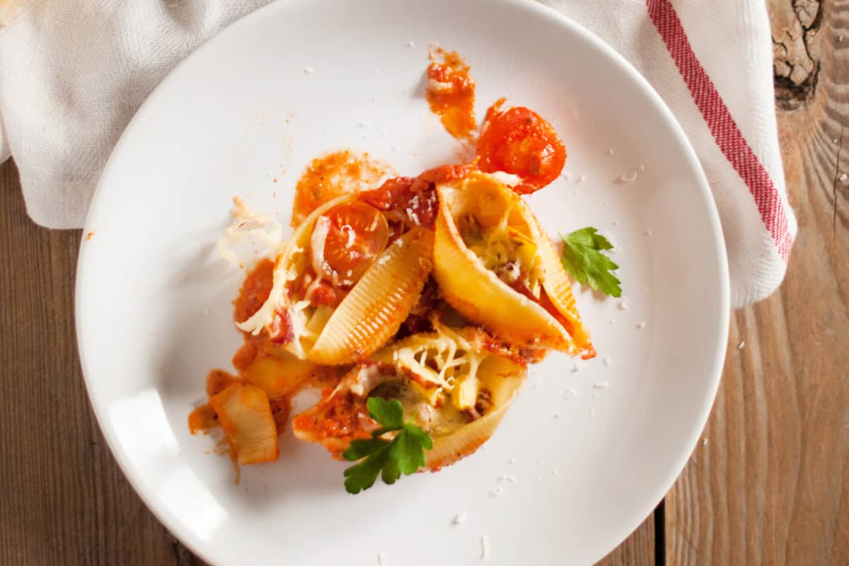 A white plate with stuffed pasta shells in tomato sauce, topped with grated cheese and garnished with parsley, sits on a wooden table next to a white napkin with a red stripe.