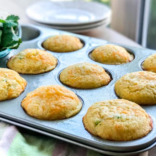 A muffin tin filled with nine golden, freshly baked cornbread muffins sits on a table beside a bowl of cilantro and stacked white plates, with sunlight streaming through a nearby window.