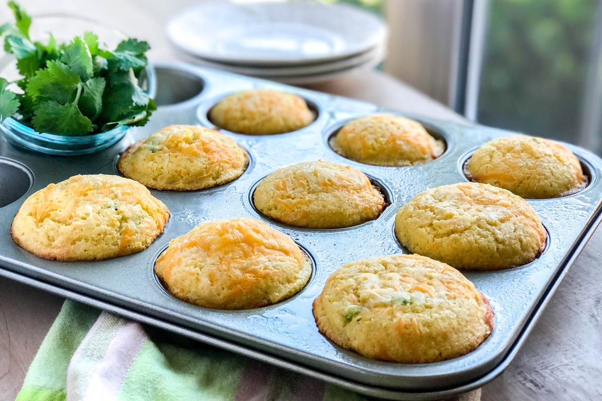 A muffin tin filled with nine golden, freshly baked cornbread muffins sits on a table beside a bowl of cilantro and stacked white plates, with sunlight streaming through a nearby window.