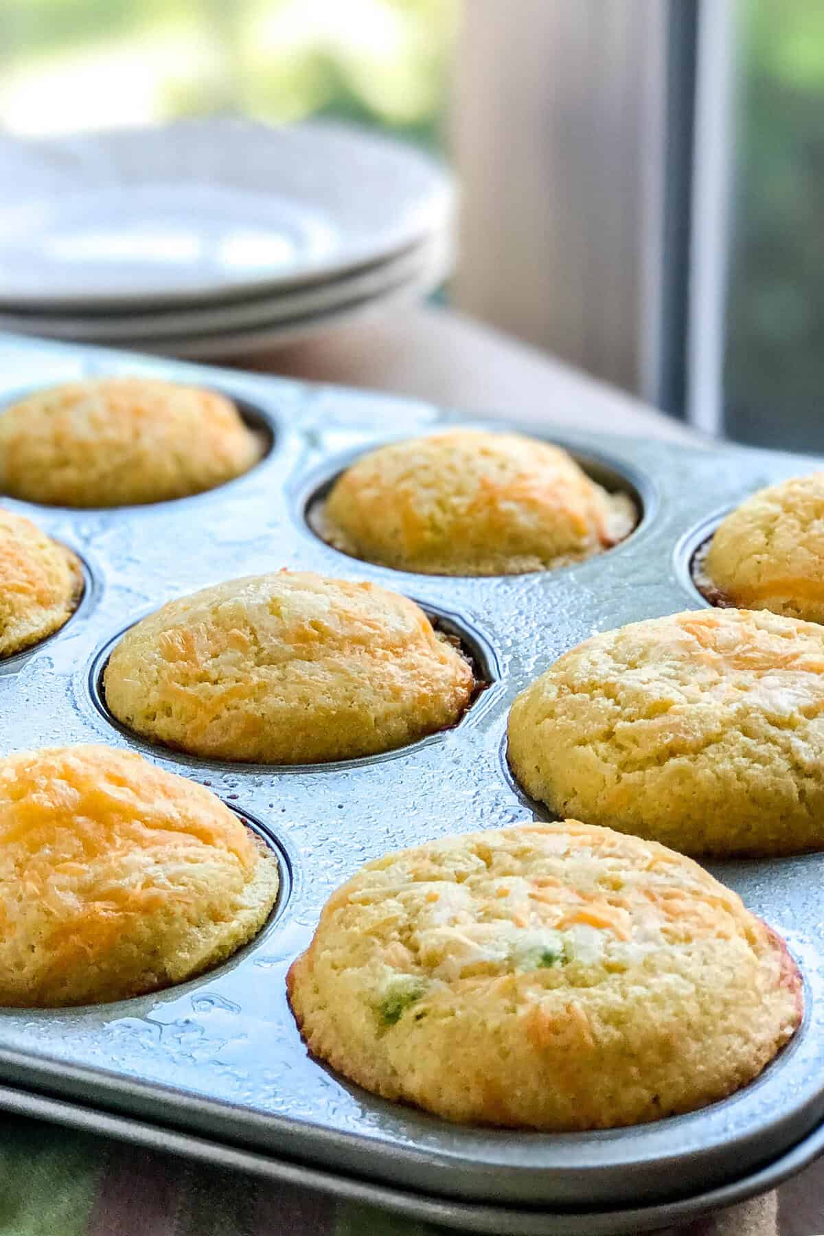 Freshly baked muffins in a muffin tin, golden brown on top, with plates stacked in the background and sunlight streaming through a nearby window.