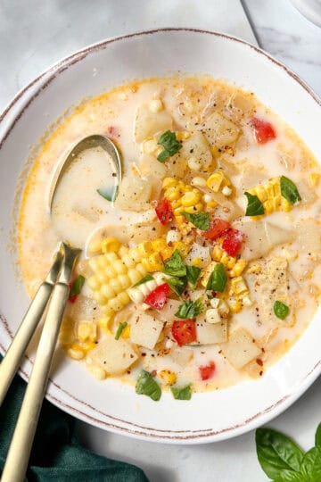 Bowl of Corn Chowder with garnishes or herbs and fresh corn with two spoons in the bowl.