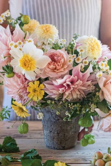 A person arranges a bouquet of pink, yellow, and white flowers in a stone vase on a rustic wooden table, creating the perfect centerpiece for a summer afternoon tea.
