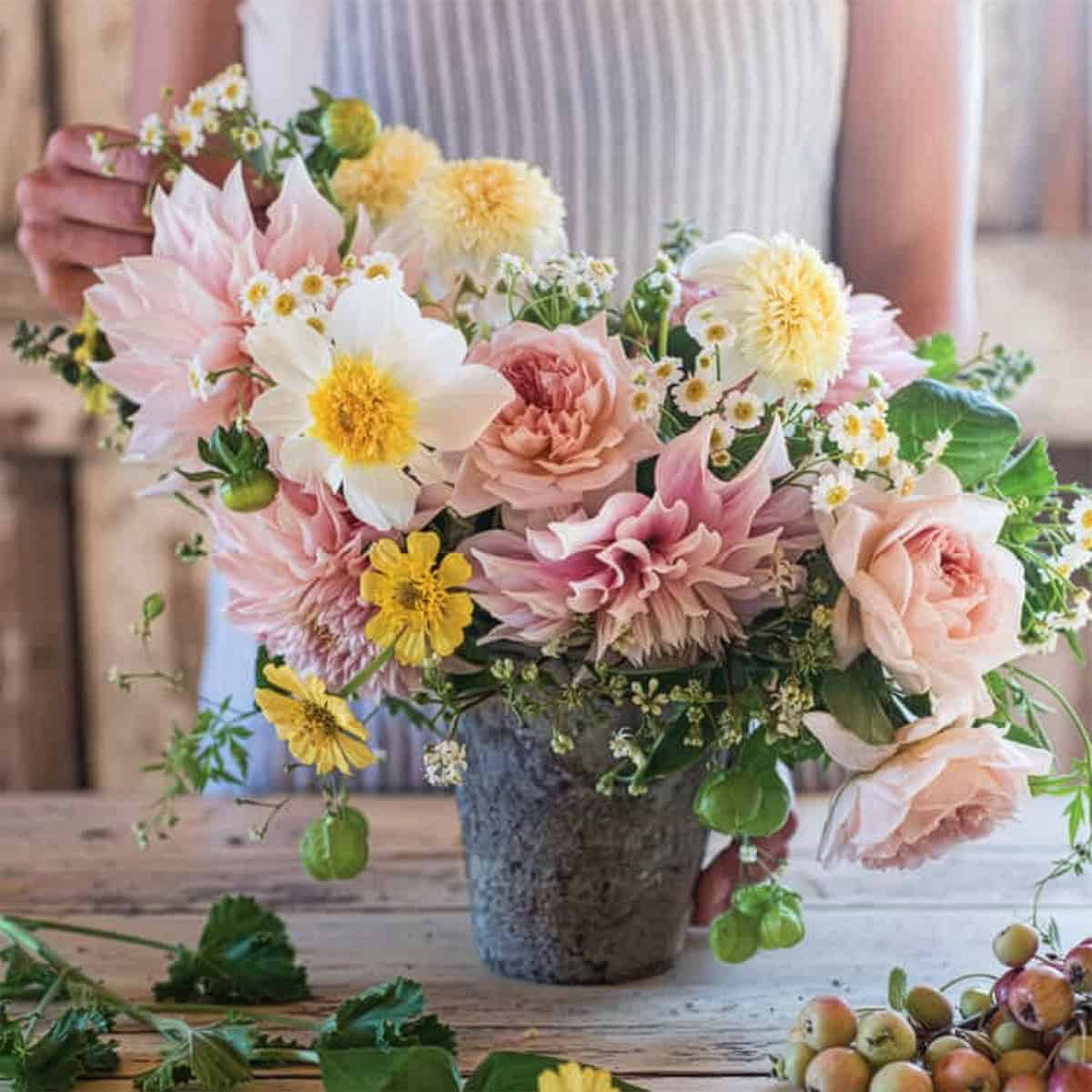 A person arranges a bouquet of pink, yellow, and white flowers in a stone vase on a rustic wooden table, creating the perfect centerpiece for a summer afternoon tea.