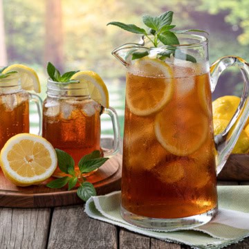 A glass pitcher and two jars of sweet tea with lemon slices and mint leaves on a table; fresh lemons and greenery in the background.