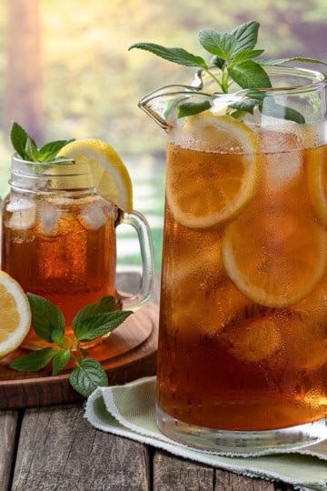 A glass pitcher and two jars of sweet tea with lemon slices and mint leaves on a table; fresh lemons and greenery in the background.