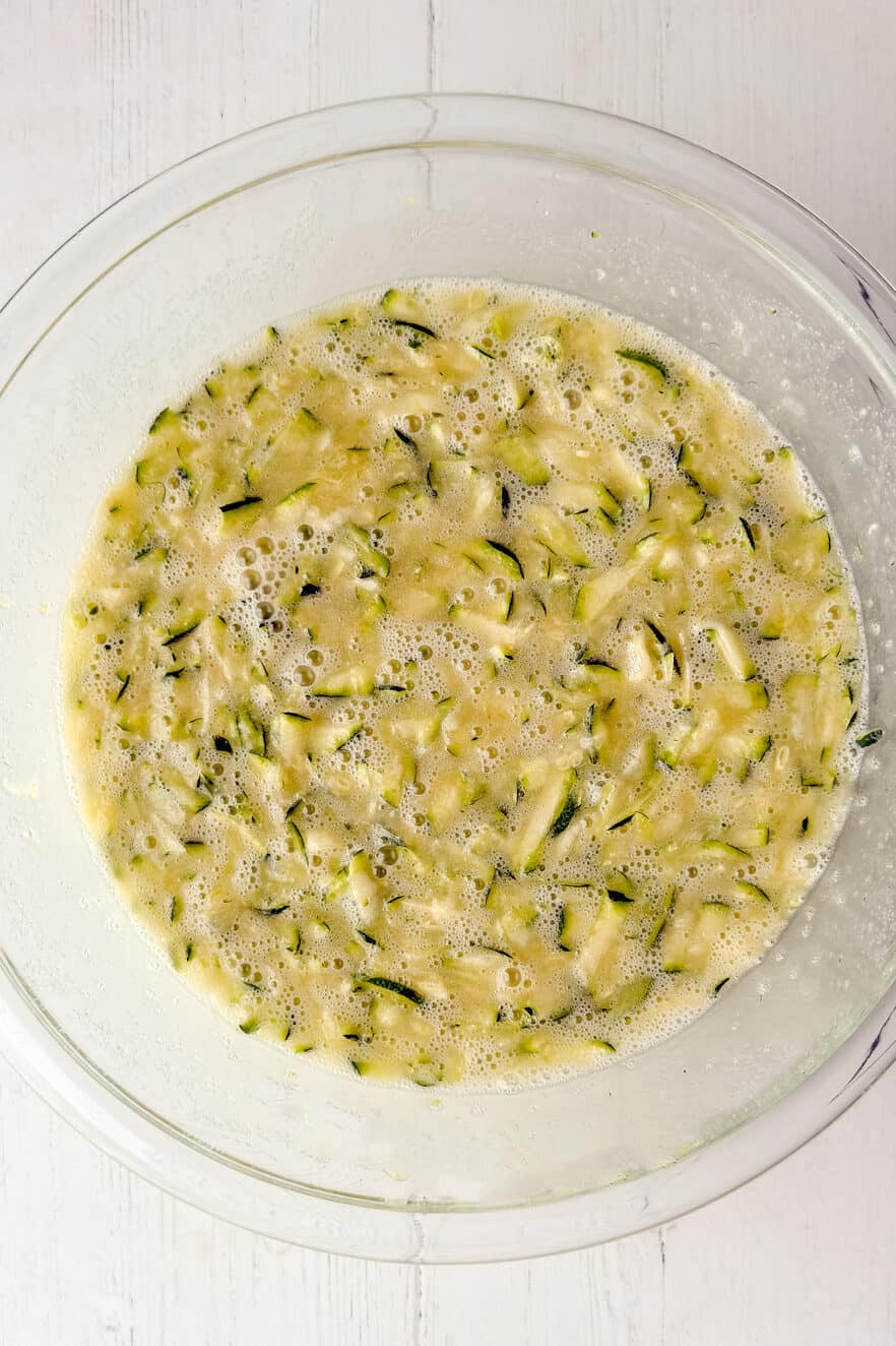 A glass bowl filled with a mixture of shredded zucchini, eggs, and liquid ingredients sits on a white wooden surface, ready to be transformed into delicious Zucchini Bread.