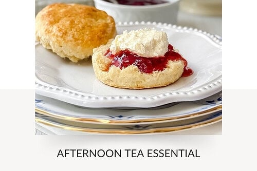 A plate with a scone topped with jam and homemade clotted cream sits in the foreground, with more scones, jam, and clotted cream visible in the background.