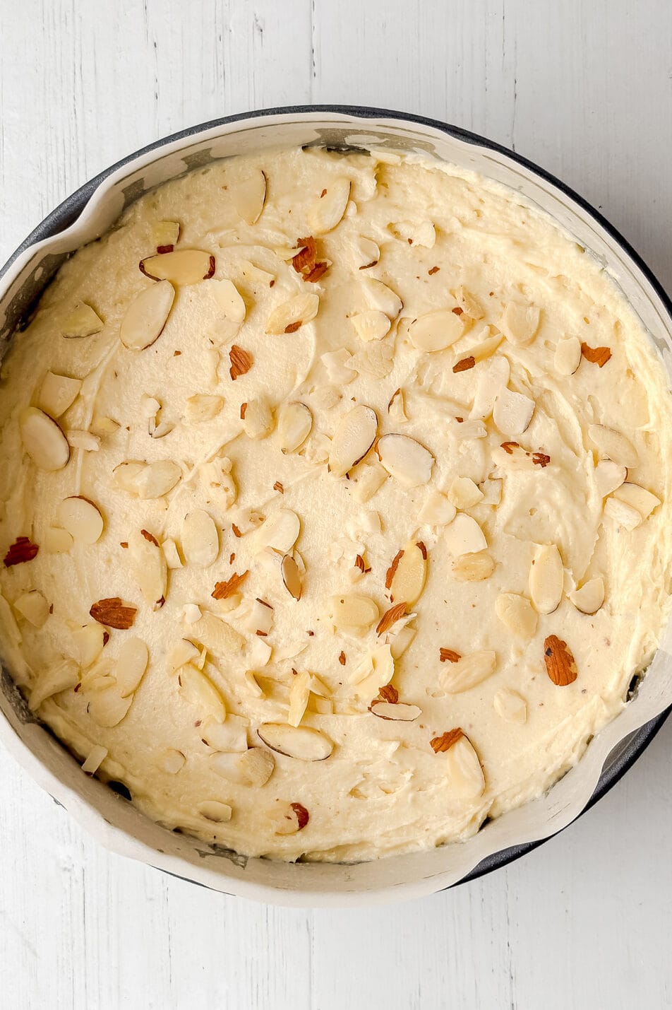 Round baking pan filled with unbaked Almond Cake batter, topped with sliced almonds, placed on a white wooden surface.
