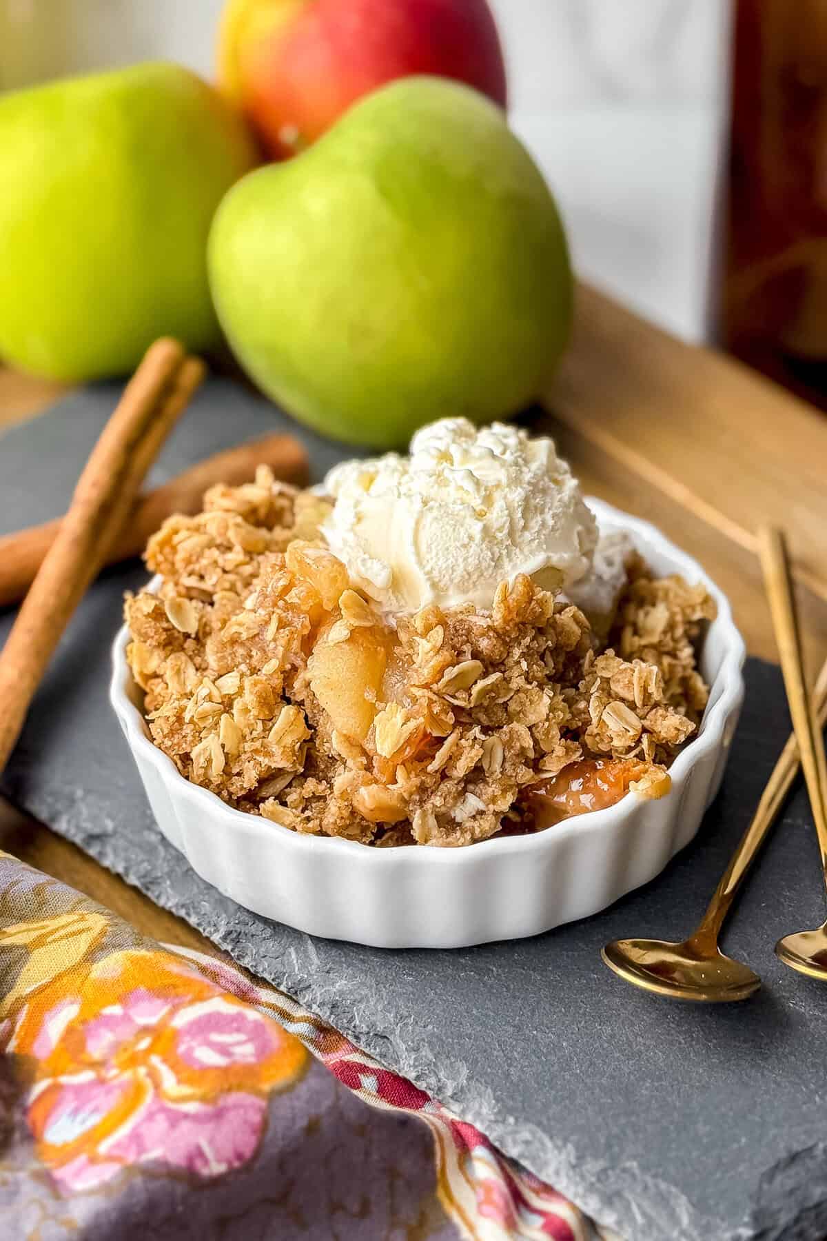 A serving of slow cooker apple crisp topped with vanilla ice cream in a white dish, with green apples, cinnamon sticks, and gold spoons in the background.
