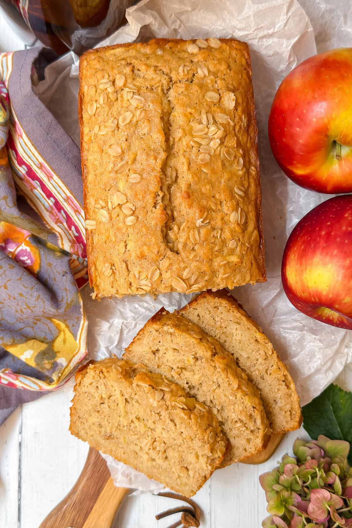 A loaf of apple oatmeal bread topped with oats sits on parchment paper, three slices cut, alongside two red apples.