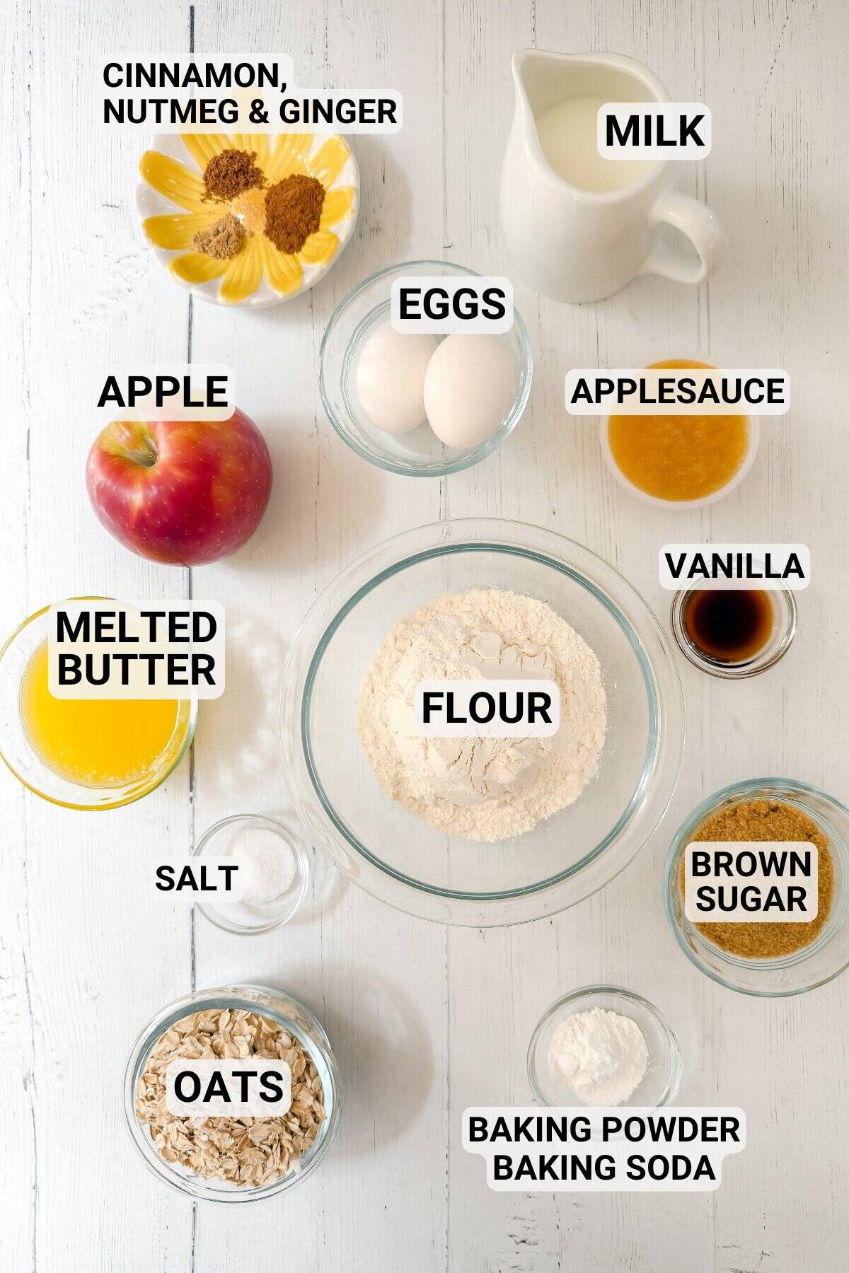 Baking ingredients on a white surface, ready to make apple oatmeal bread: flour, oats, apple, applesauce, eggs, milk, melted butter, brown sugar, vanilla, spices, salt, baking powder, and baking soda.
