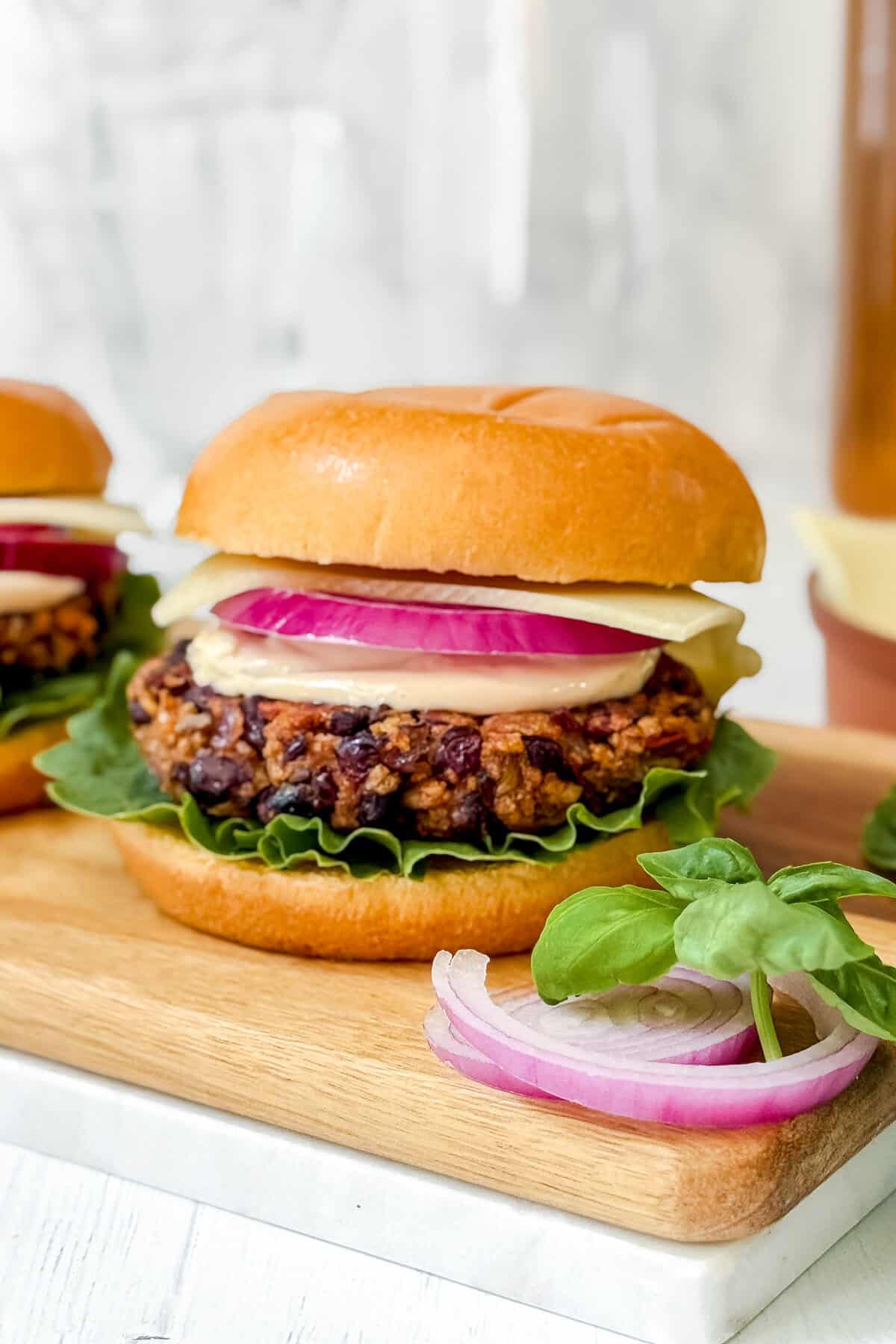 A black bean veggie burger with lettuce, a thick slice of red onion, and cheese, served on a bun. The burger sits on a wooden board with fresh basil and sliced red onion beside it.