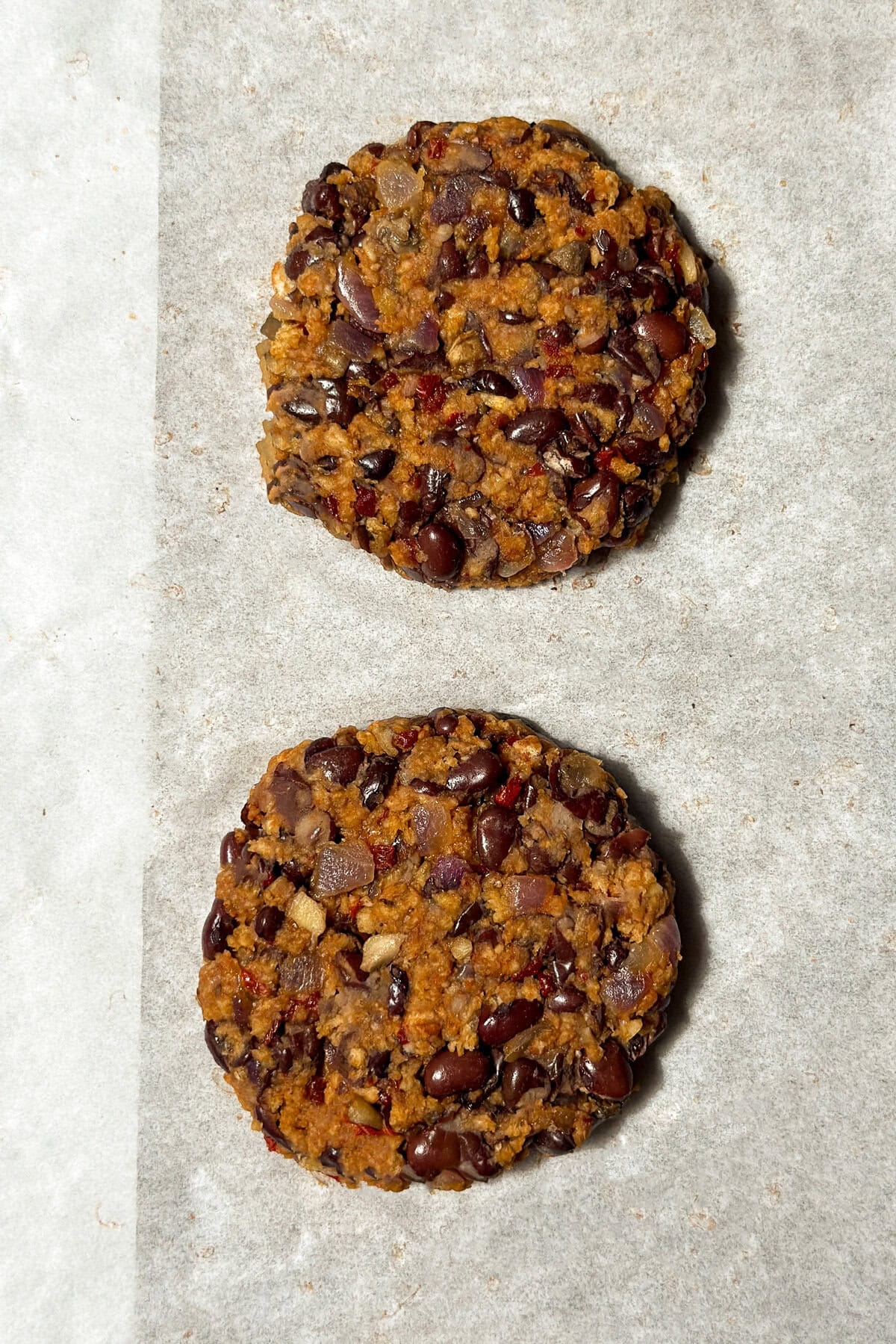 Two homemade black bean burger patties on a sheet of parchment paper, spaced apart. The patties have a textured surface with visible beans and chopped vegetables.