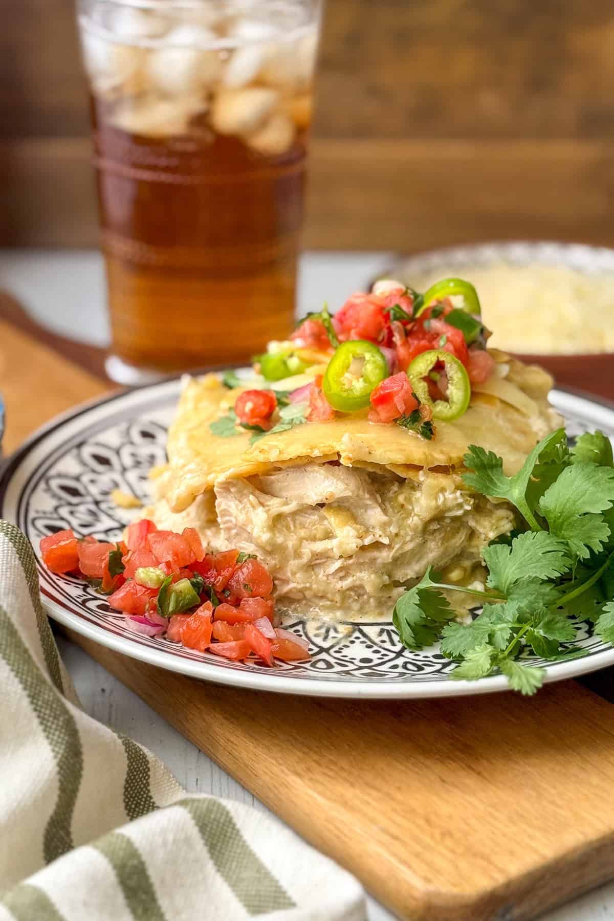 A slice of creamy crock pot chicken enchilada casserole topped with salsa and jalapeños, served with fresh cilantro and a glass of iced tea in the background.