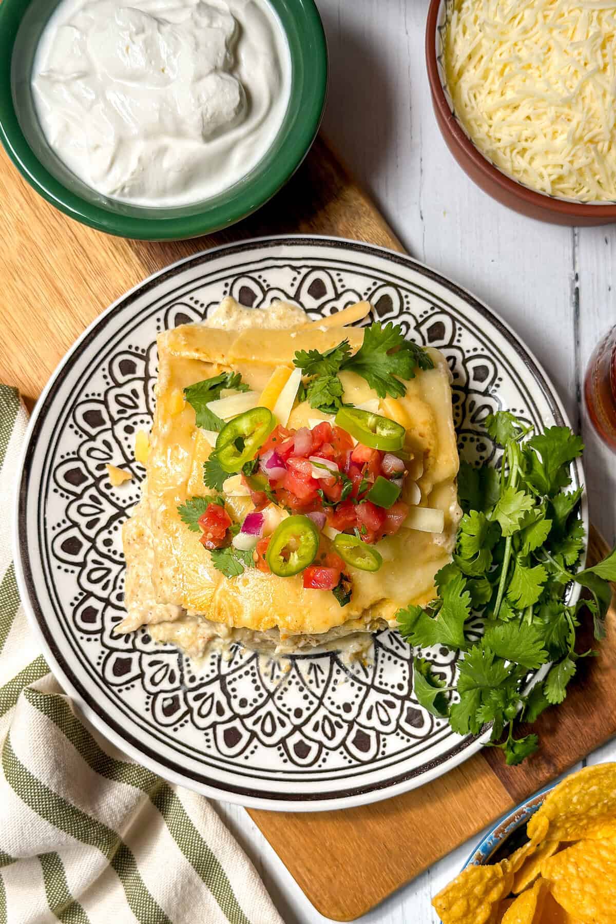 A plate of cheesy crock pot chicken enchilada casserole topped with diced tomatoes, jalapeños, and cilantro, served with fresh cilantro on the side; bowls of sour cream and shredded cheese nearby.