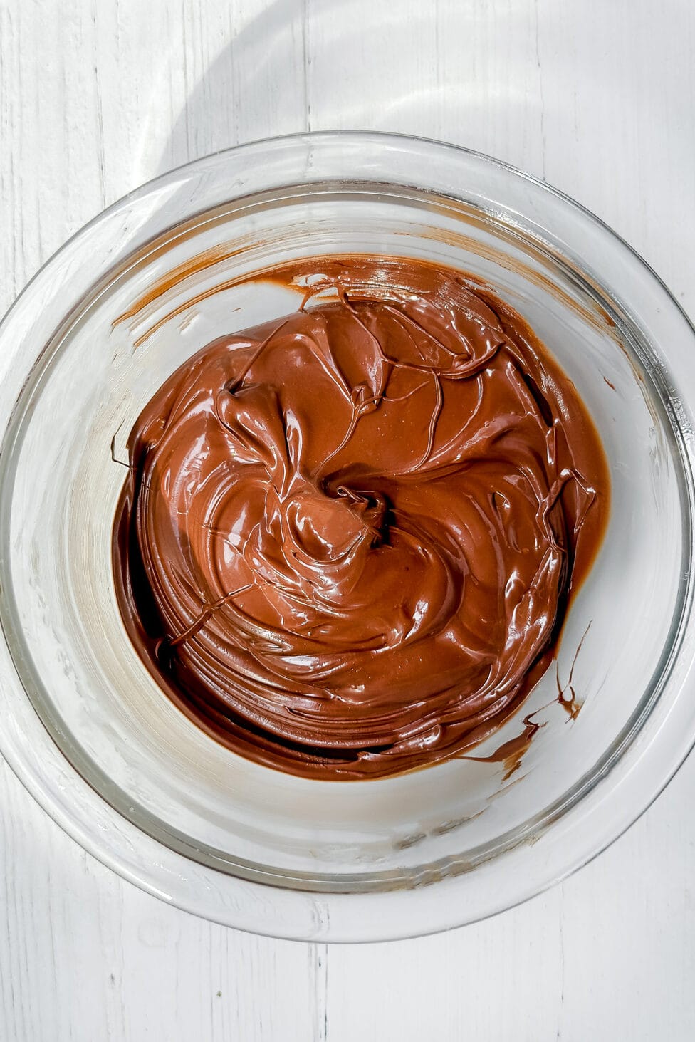 A glass bowl filled with smooth, melted chocolate for a delicious chocolate pie sits on a white wooden surface.