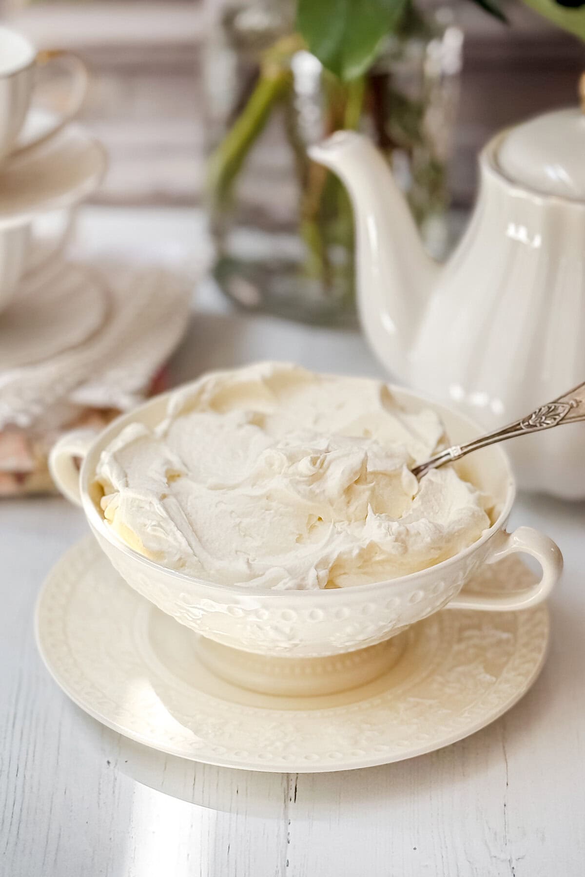 A bowl of homemade clotted cream, made with just 3 ingredients, sits with a spoon on a saucer next to a white teapot and stacked teacups.