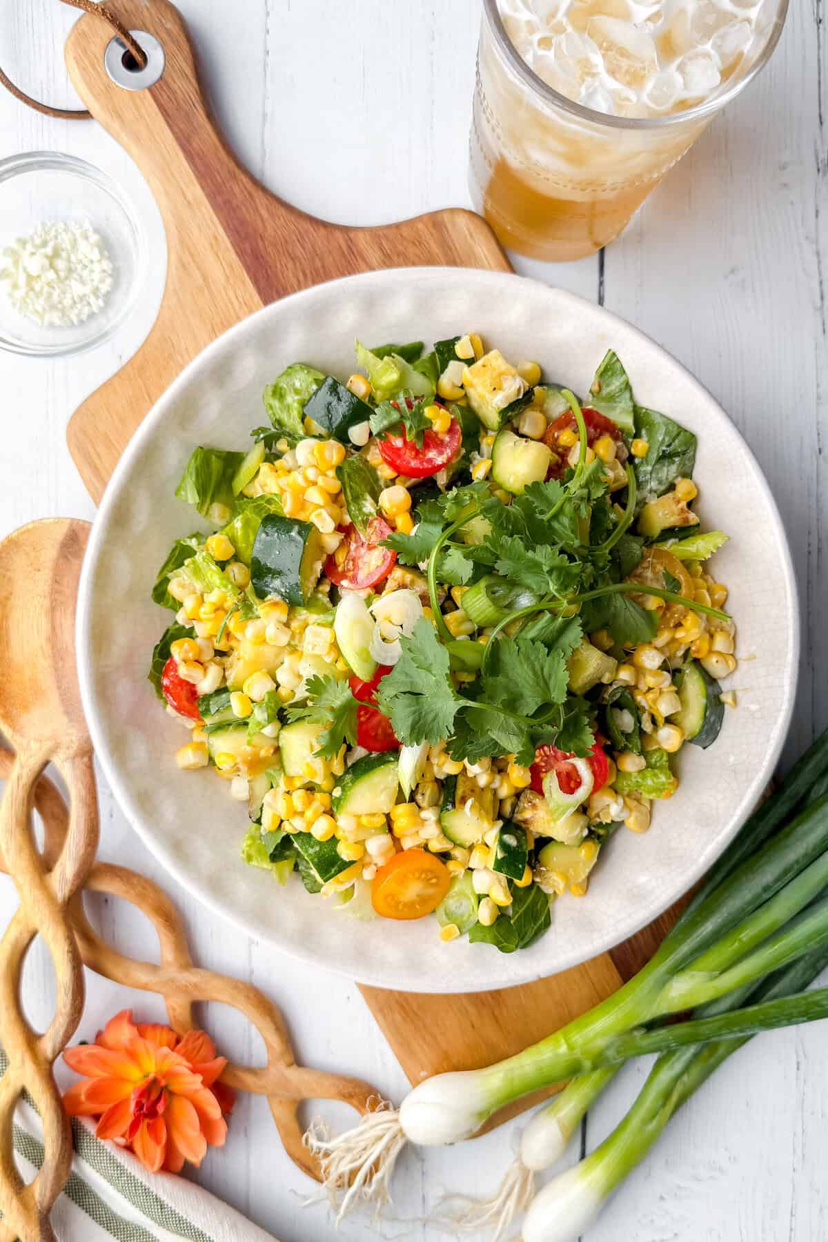 A bowl of fresh Corn Salad with cherry tomatoes, cucumber, spinach, cilantro, and corn sits on a white table with green onions, a drink, and a cutting board nearby.