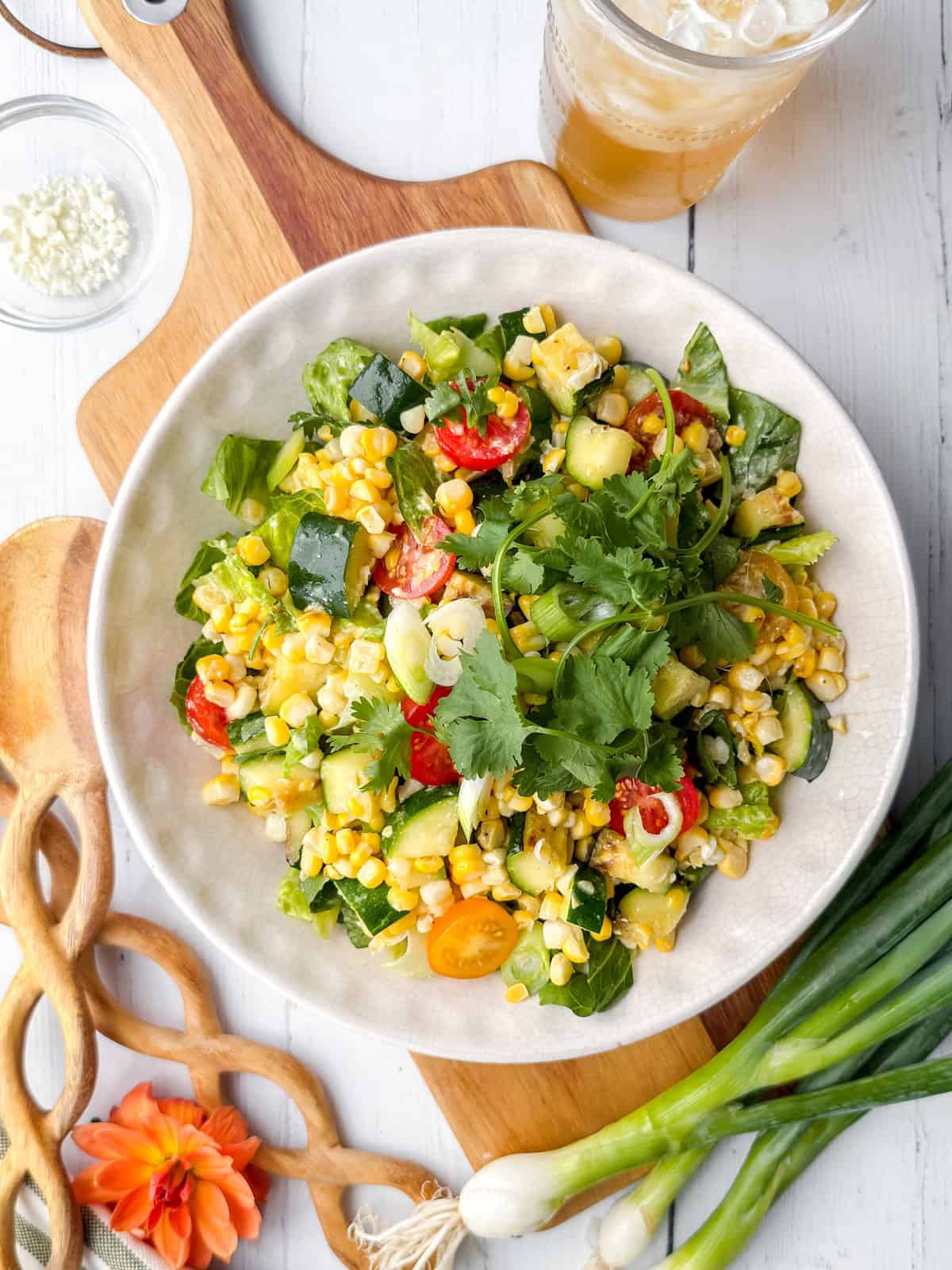 A bowl of fresh grilled corn zucchini salad with tomatoes, cucumber, and cilantro sits on a light table, next to green onions, an iced drink, and a cutting board.