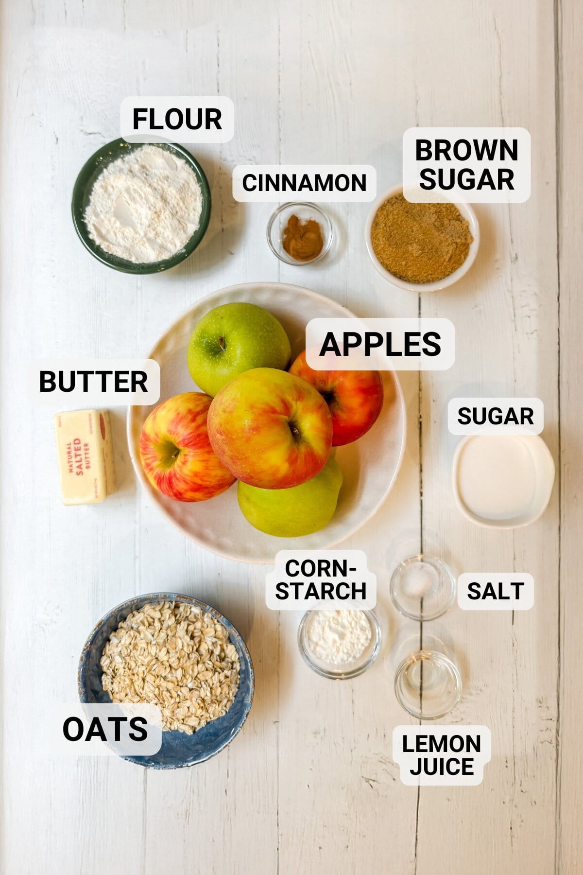 A variety of ingredients for slow cooker apple crisp on a white table, including apples, flour, oats, butter, brown sugar, sugar, cinnamon, cornstarch, salt, and lemon juice-each carefully labeled.