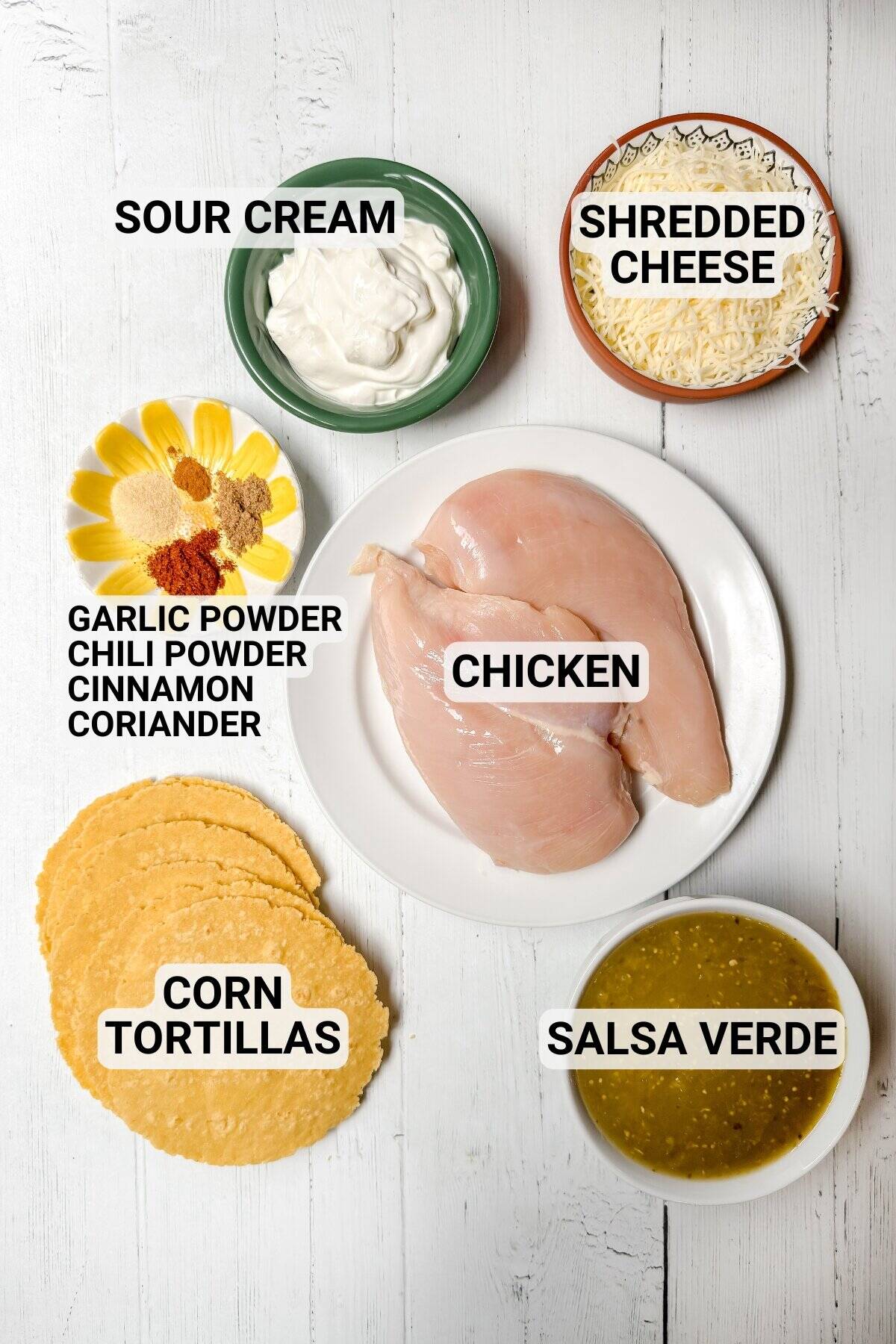 Ingredients for a Chicken Enchilada casserole are laid out on a white surface, featuring raw chicken breasts, sour cream, shredded cheese, spices, corn tortillas, and salsa verde-perfect for an easy crock pot meal.