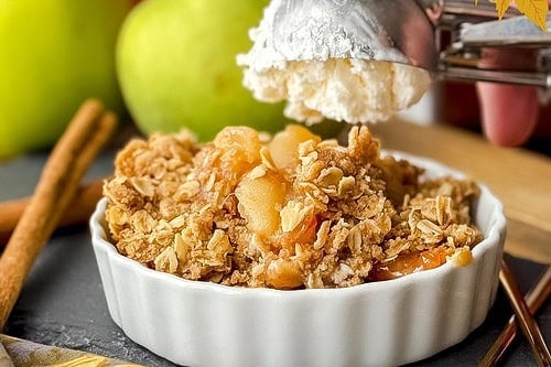 A serving of slow cooker apple crisp with a scoop of ice cream in a bowl, sliced apples in a crockpot below, and decorative fall leaves. Text reads "Crockpot Apple Crisp.