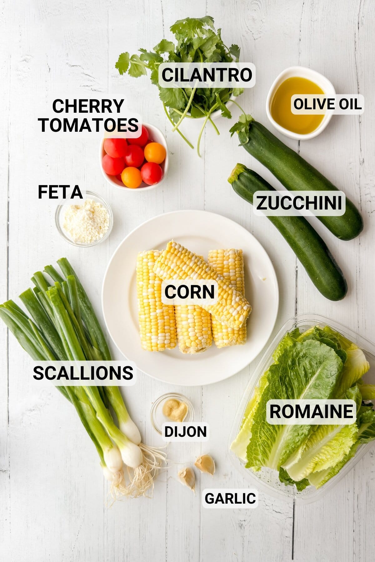 Top-down view of common salad ingredients on a white table, including grilled zucchini, corn, romaine, cherry tomatoes, feta, scallions, cilantro, olive oil, garlic, and Dijon mustard-perfect for a fresh grilled zucchini salad or corn salad.