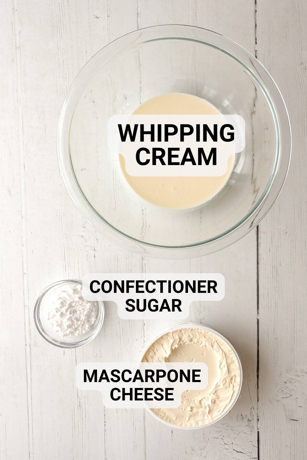 A glass bowl of whipping cream, a small bowl of confectioner sugar, and a bowl of mascarpone cheese on a white wooden surface-perfect for making an easy 3 Ingredient Clotted Cream. Each ingredient is clearly labeled.