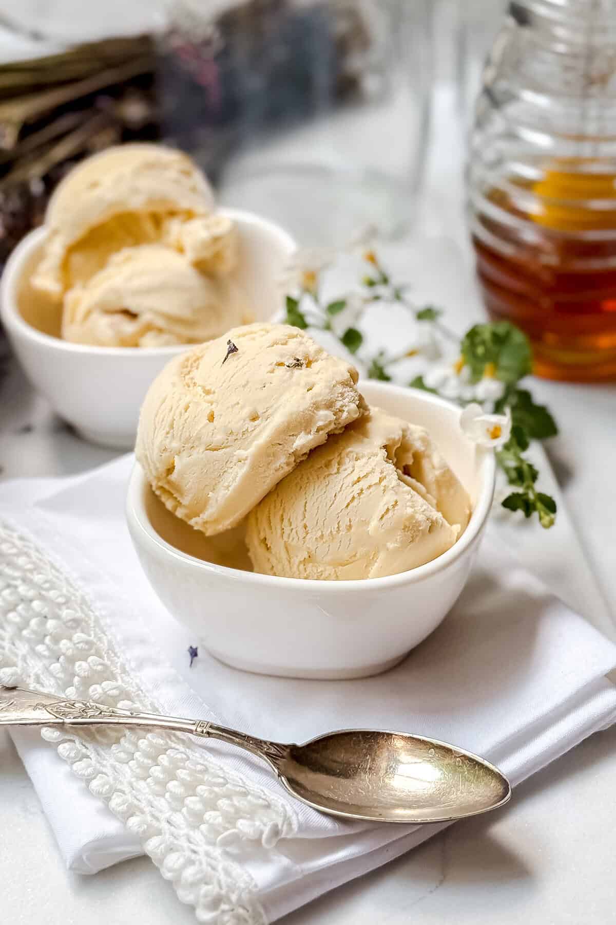 Two white bowls filled with scoops of creamy honey lavender ice cream sit on a white napkin beside a silver spoon. A glass jar of honey and small white flowers are in the background.