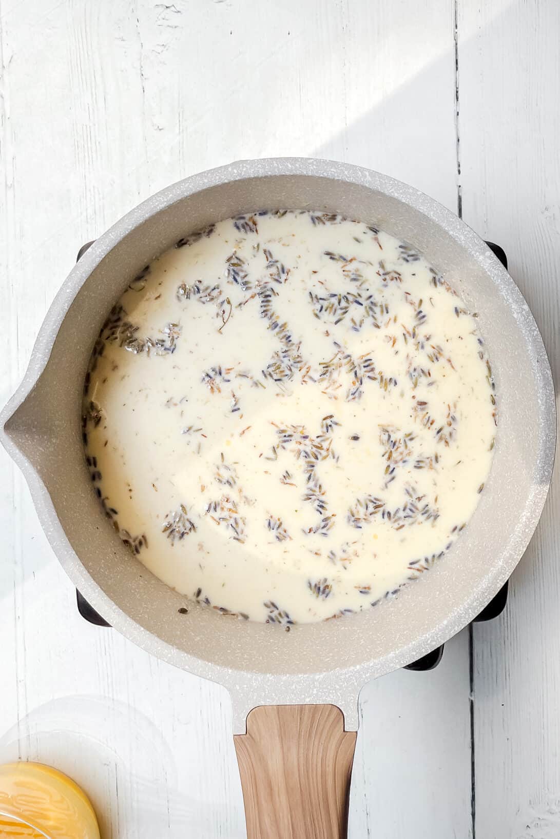 A saucepan with a wooden handle sits on a white surface, filled with cream and dried lavender buds. A small jar of honey is partially visible in the bottom left corner.