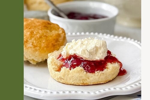 A scone topped with jam and homemade clotted cream sits on a white plate, with more scones and a bowl of jam in the background. Text reads, “Mock Clotted Cream for Tea Scones! 3 Ingredients.”.