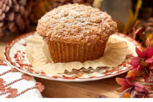 A pumpkin muffin dusted with powdered sugar sits on a decorative plate, surrounded by autumn-themed decorations, with another in the background. Text reads: "Panera Pumpkin Muffins easy copycat recipe.