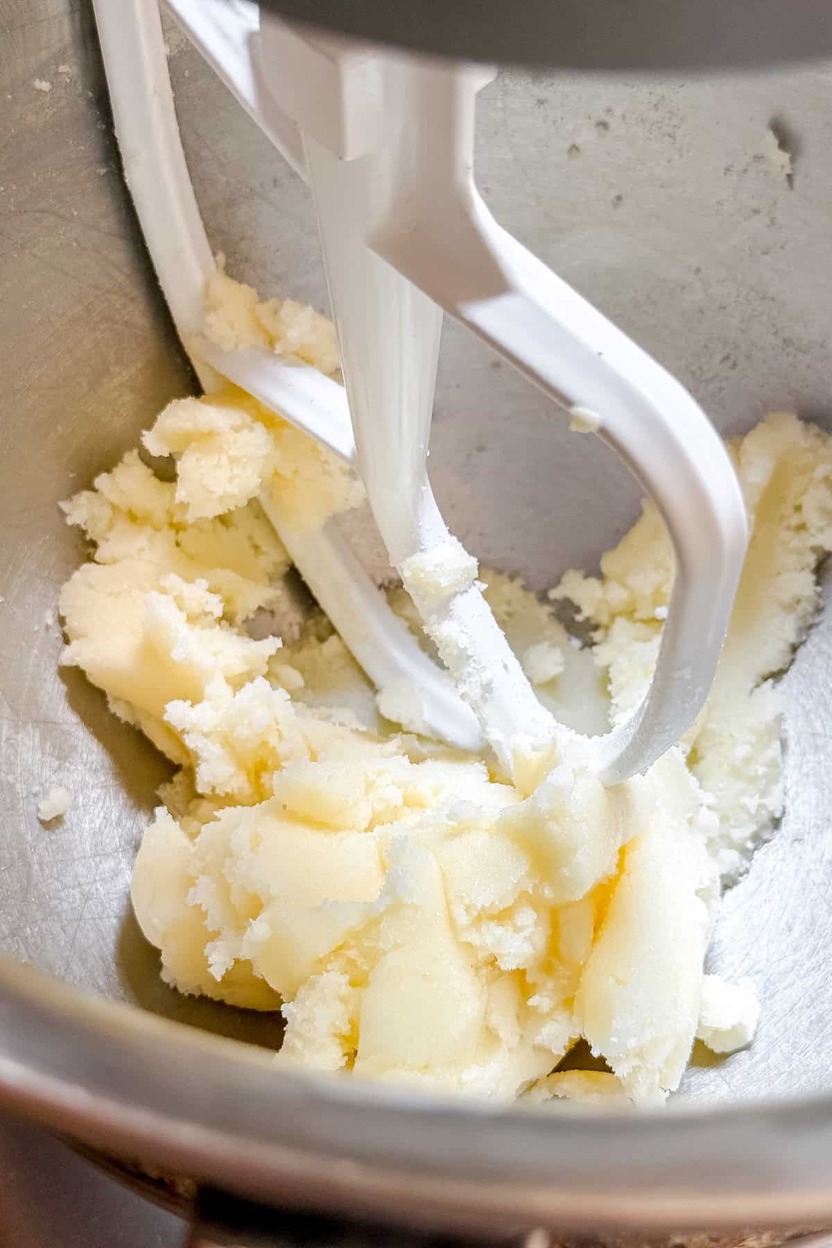 A close-up of butter and sugar being creamed together in a metal mixing bowl with a stand mixer paddle attachment, preparing the base for a delicious peach cobbler cake.