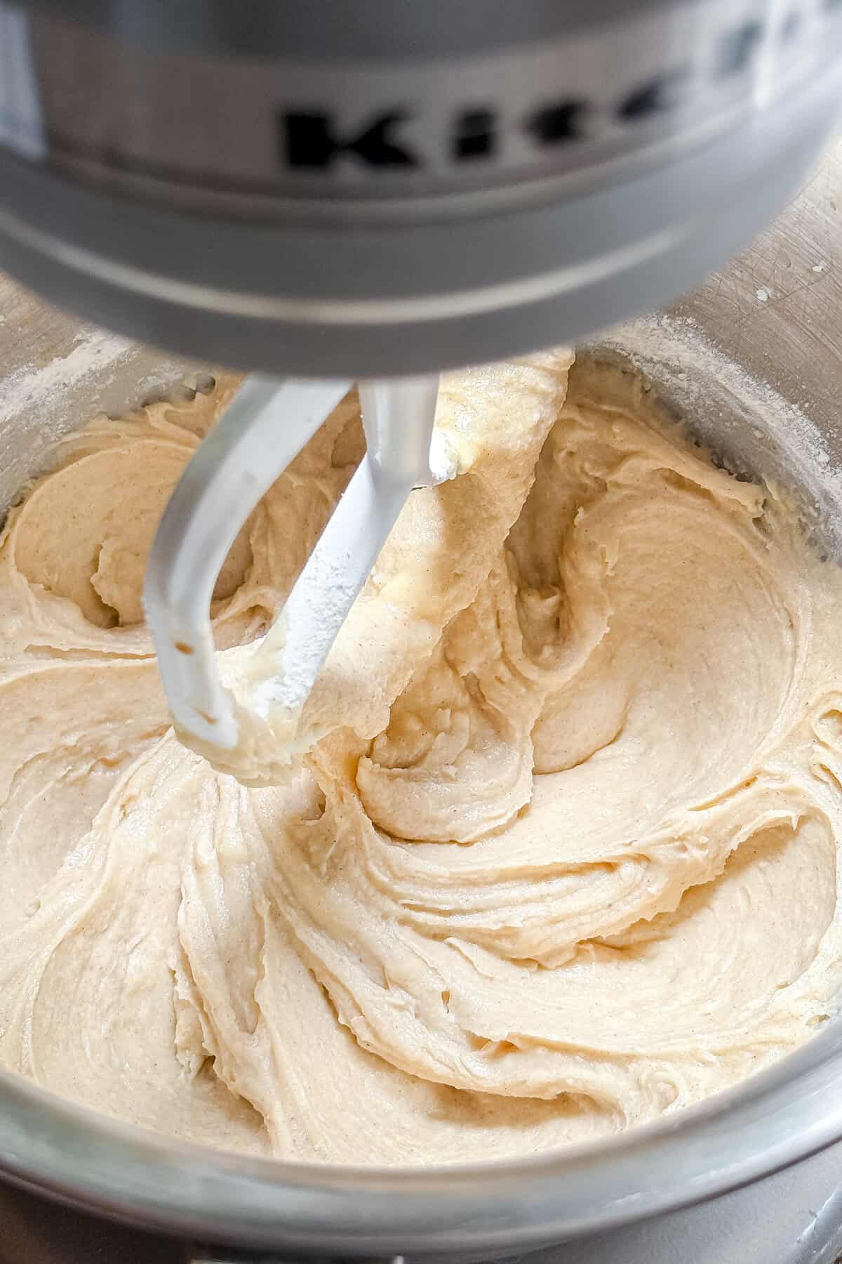 Close-up of a stand mixer blending a creamy, light-colored peach cobbler cake batter in a metal mixing bowl with a paddle attachment.