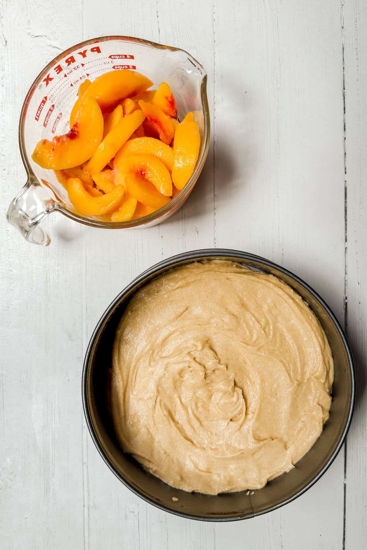 A glass measuring cup with sliced peaches, perfect for peach cobbler cake, sits next to a round cake pan filled with unbaked batter on a white wooden surface.