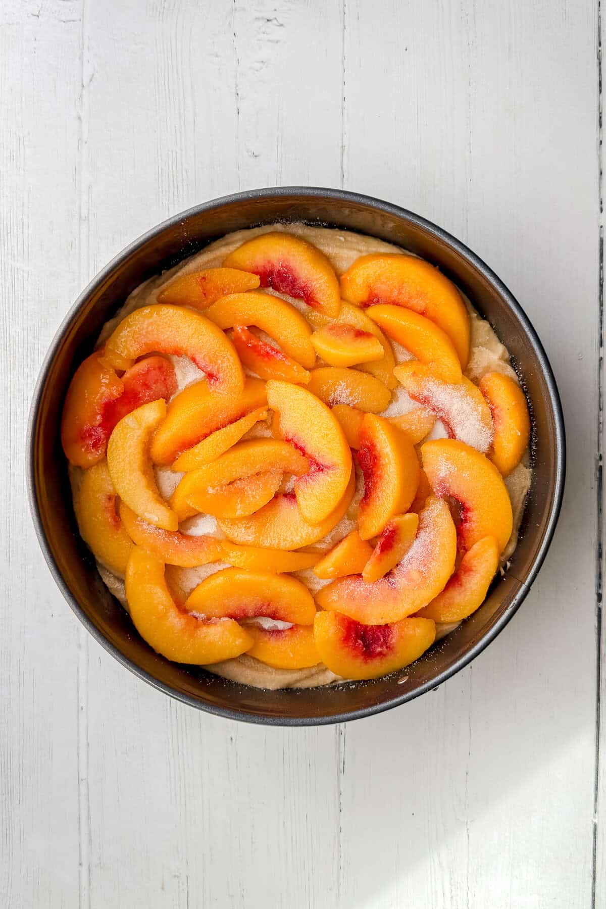 A round baking pan filled with a layer of sliced peaches arranged on top of an unbaked peach cobbler cake batter, sprinkled with sugar, sits on a white wooden surface.