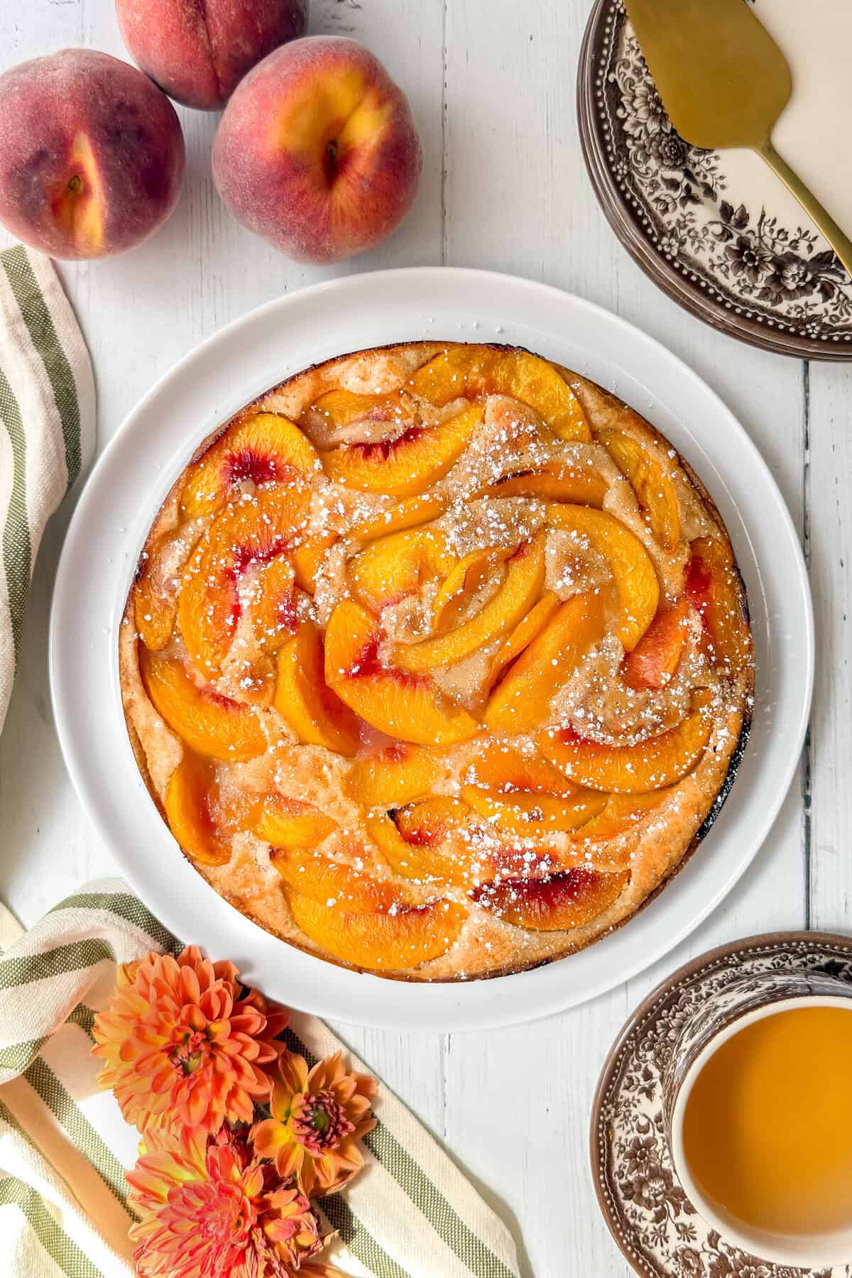 A round peach cobbler cake dusted with powdered sugar sits on a white plate. Nearby are fresh peaches, orange flowers, a striped cloth, a cup of tea, and a patterned plate with a pie server on a white wooden surface.