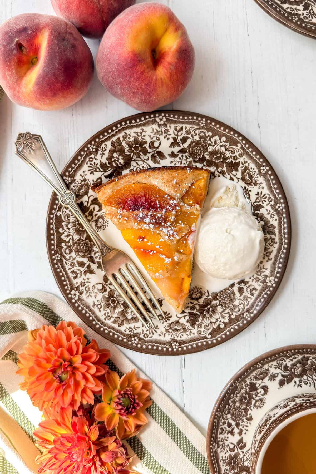 A slice of peach cobbler cake with a scoop of vanilla ice cream rests on a vintage brown floral plate, surrounded by fresh peaches, orange flowers, and a cup of tea on a white table.